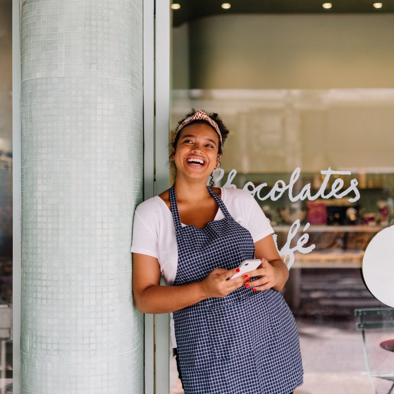 Successful cafe owner, a young woman, stands in her restaurant using a smartphone. Happy female entrepreneur using technology to manage her small business efficiently.